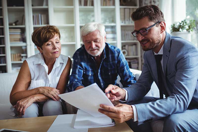 A man in a suit shows documents to an older couple seated on a couch, likely discussing retirement planning or other financial matters in a home office setting.